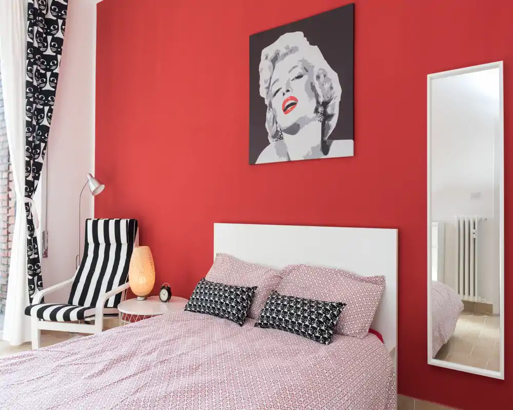 Stylish bedroom with a patterned pink duvet, two decorative cushions and a white headboard set against a bold red accent wall featuring pop art. Natural light brightens the space; a striped armchair and bedside lamp add comfort and character.