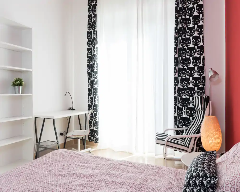 Bright, modern bedroom with patterned textiles and a comfortable bed in the foreground, large window with sheer curtains, a small desk and a striped armchair.