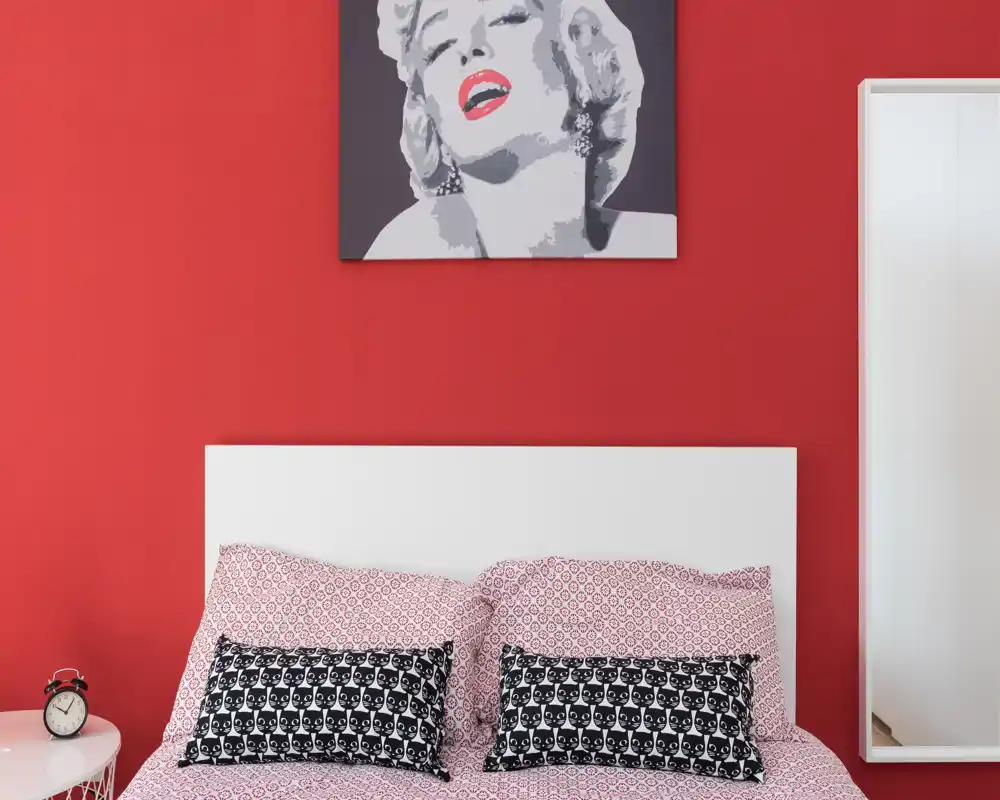 Bright, stylized bedroom with a white headboard, patterned bedding and decorative cushions against a bold red wall featuring pop-art above the bed.