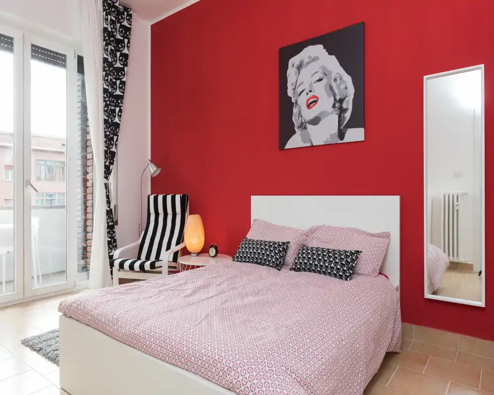 Bright, stylish bedroom with a large double bed against a red accent wall, patterned duvet, bedside lamp and a comfortable striped armchair by the balcony doors.