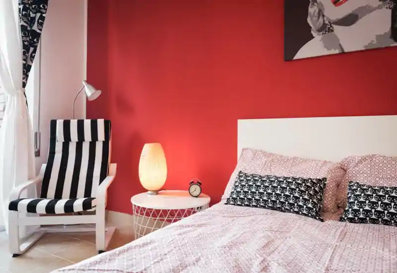 Bright, stylish bedroom with a patterned duvet, decorative pillows and a bedside lamp. The bold red accent wall and coordinated black-and-white chair create a modern, cozy atmosphere.