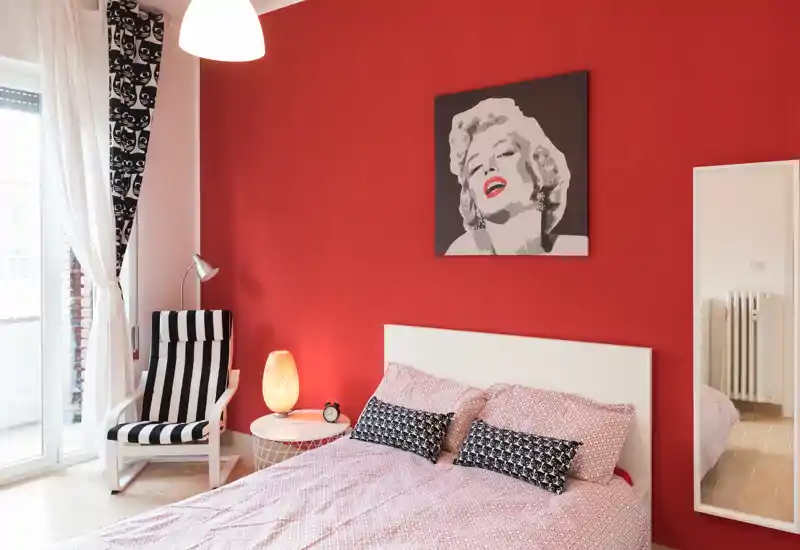 Bright, stylish bedroom with a full bed against a bold red feature wall, decorative cushions, a reading chair by the window and modern artwork above the headboard.