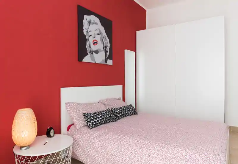 Bright, stylish bedroom with a queen bed dressed in patterned linens, a white headboard, bedside table with warm lamp, and bold red accent wall featuring pop-art decor.