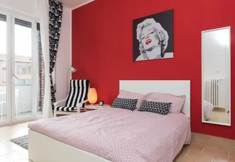Camera da letto luminosa e di stile con letto matrimoniale, cuscini decorativi, poltrona per la lettura e accesso al balcone. Parete rossa e opera d'arte moderna creano un'atmosfera accogliente e chic.