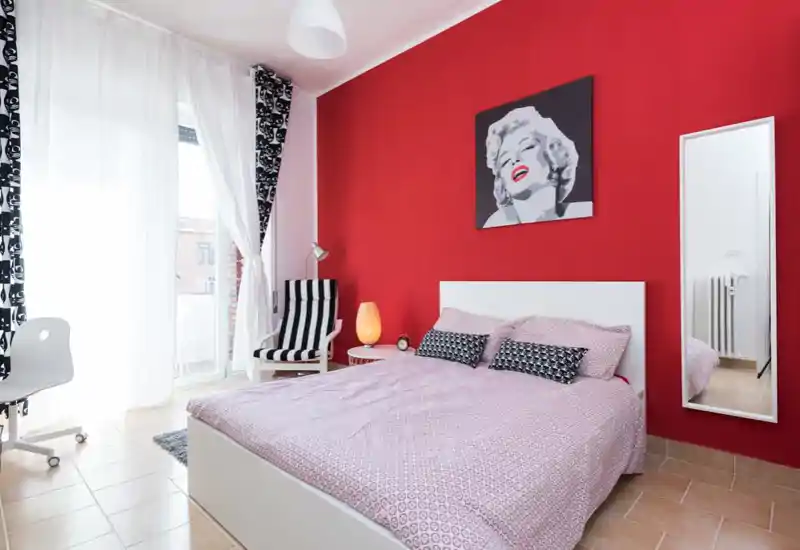Bright, stylish bedroom with a white bed, pink patterned bedding and a bold red accent wall featuring pop-art. Large balcony window provides abundant natural light; striped armchair and full-length mirror complete the modern, cozy layout.