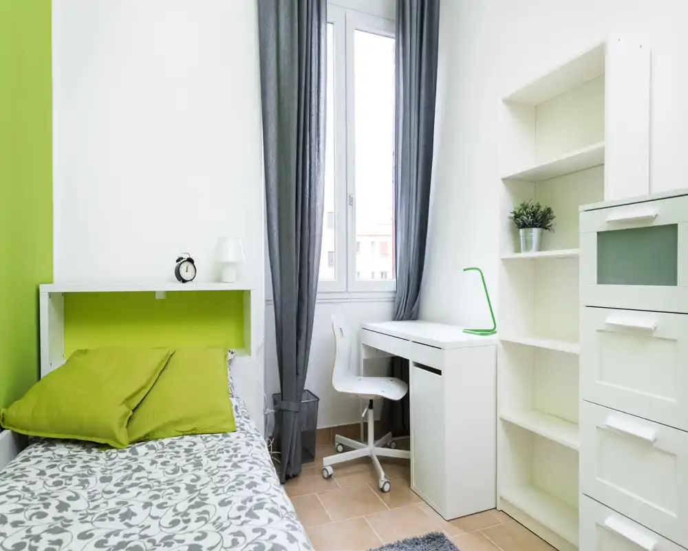 Small, bright single bedroom with a comfortable bed, white desk by the window and built-in shelving. Natural light and fresh green accents create a calm, modern feel ideal for a student or short-term stay.