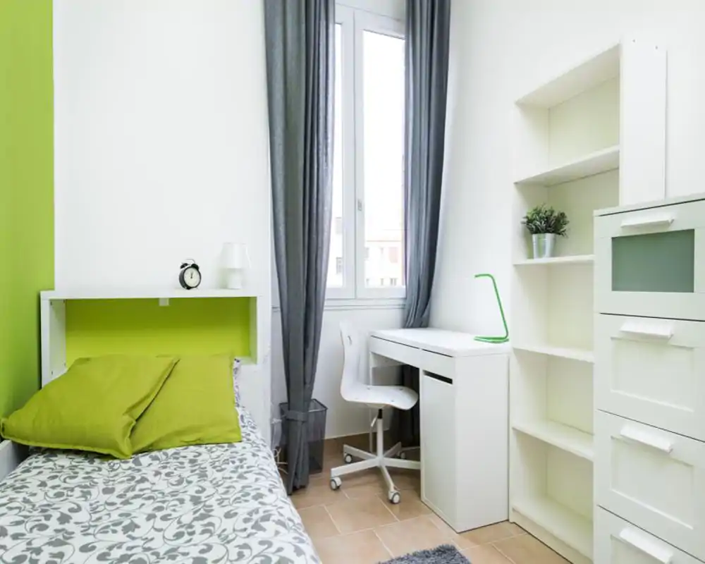 Bright, modern single bedroom with a neatly made bed, green accent wall and matching cushions, a compact desk by the window and shelving for storage — ideal for a student or single occupant.