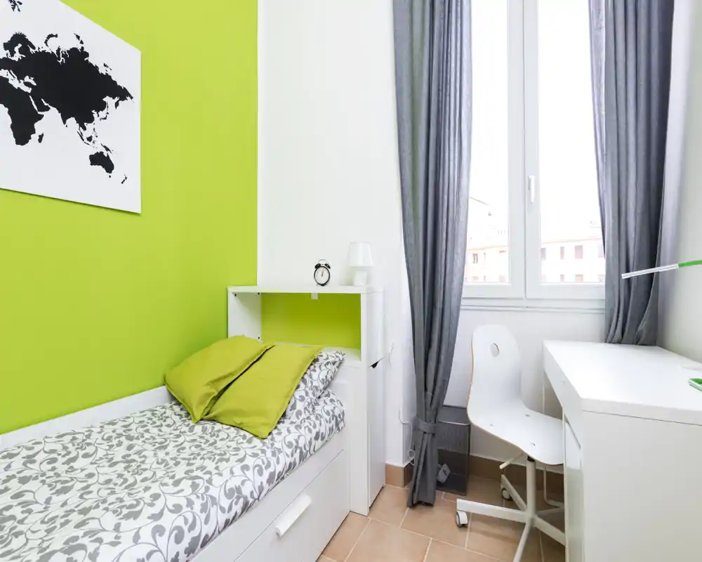 Small bright bedroom with a single bed, patterned bedding and green accent wall. A white desk and chair sit by the window offering a tidy, functional workspace.