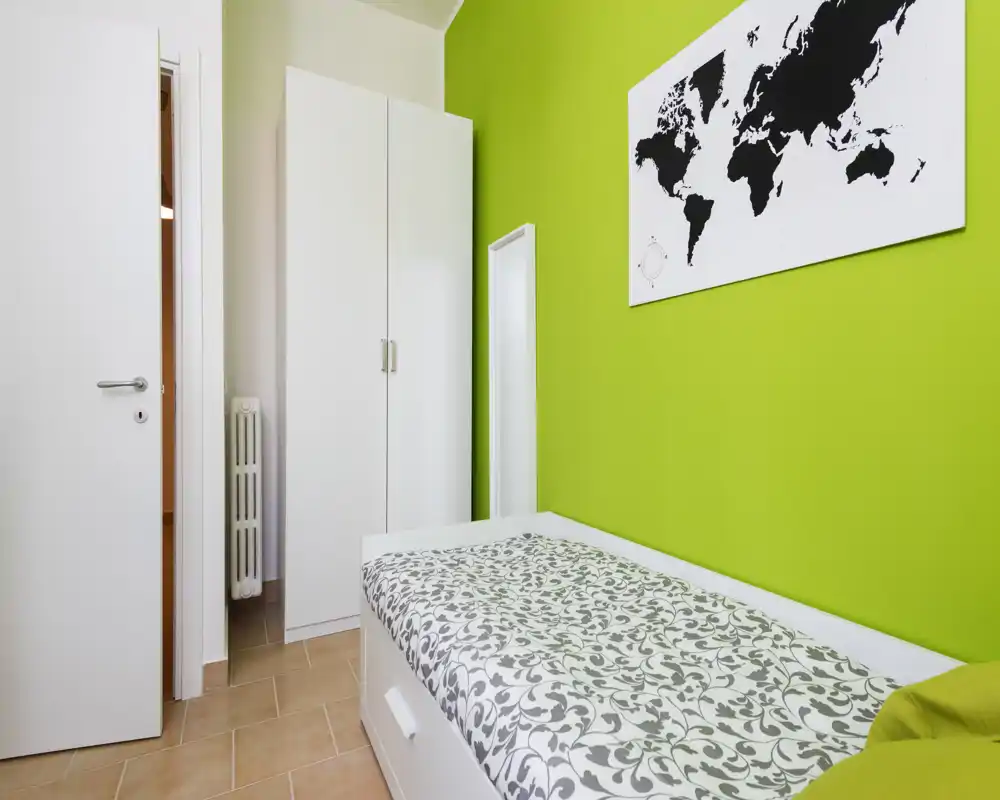 Compact single bedroom with a white bed frame, patterned bedding and a bright green accent wall; includes a tall white wardrobe and wall map decor. Bright, minimal and freshly styled — ideal for a single tenant or short stays.