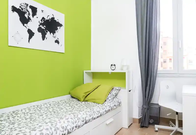 Bright, compact bedroom with a single bed dressed in patterned linens, green accent pillows and a decorative world map. Natural light from the window and a small desk make this room ideal for a student or short-term stay.