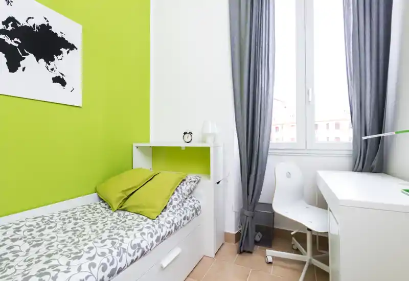 Small bright bedroom with a single bed, patterned bedding and green accent wall. A white desk and chair sit by the window offering a tidy, functional workspace.