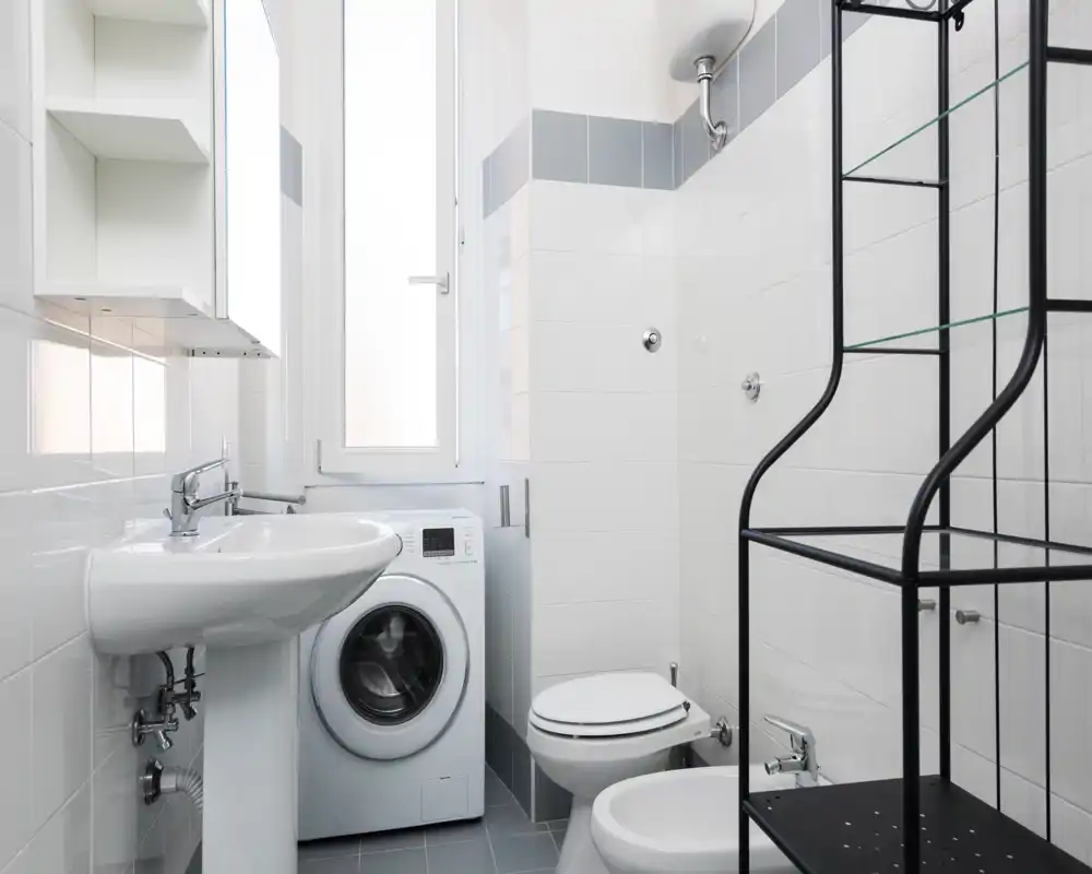 Compact, modern bathroom with white tiles, pedestal sink, toilet, bidet and a washing machine under natural window light.