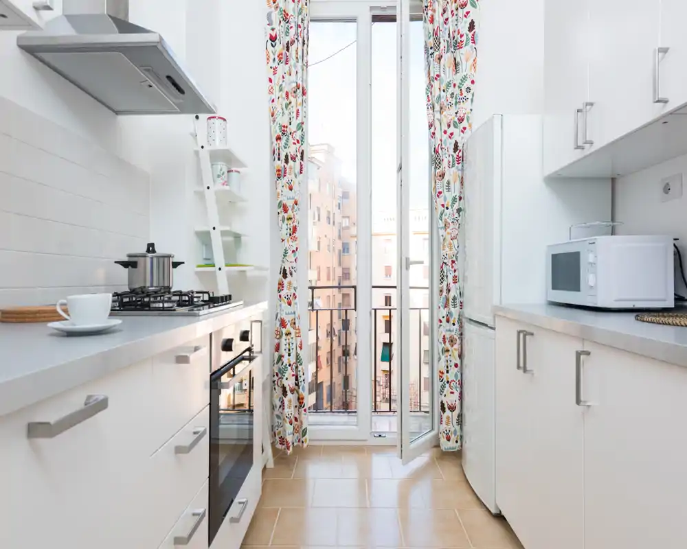 Bright, modern galley kitchen with white cabinets, gas hob, oven and a microwave. Large glass door with patterned curtains brings in abundant natural light and a small balcony view.