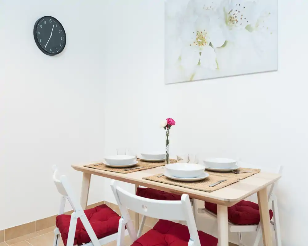 Bright, neatly set dining area with a wooden table for four, woven placemats, white dishware and red seat cushions creating a welcoming atmosphere.