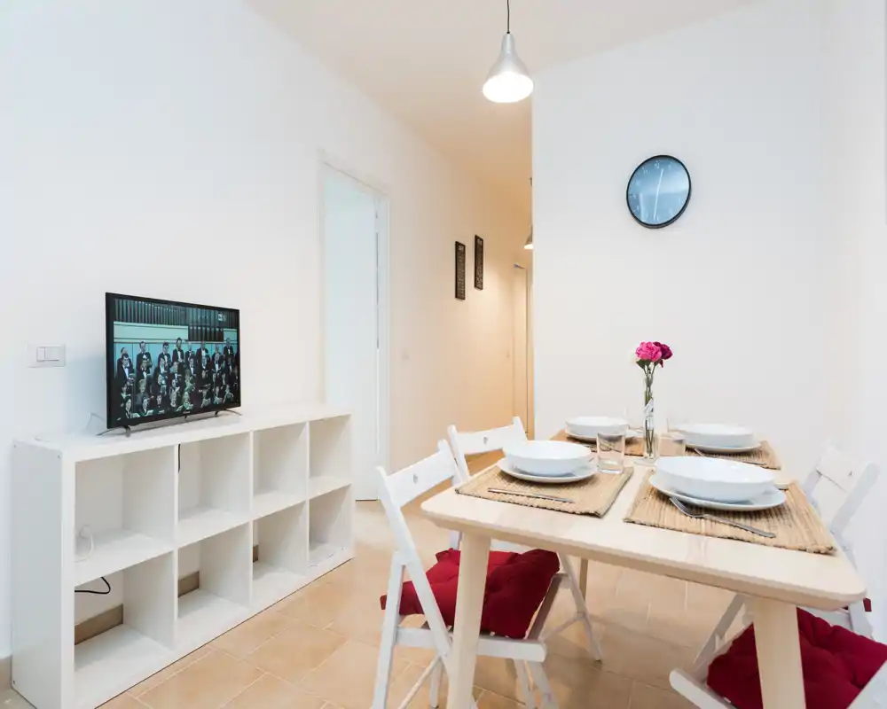 Bright, modern dining area with a wooden table set for four, woven placemats and a small flower centerpiece; simple shelving and a wall clock complete the clean, minimalist look.