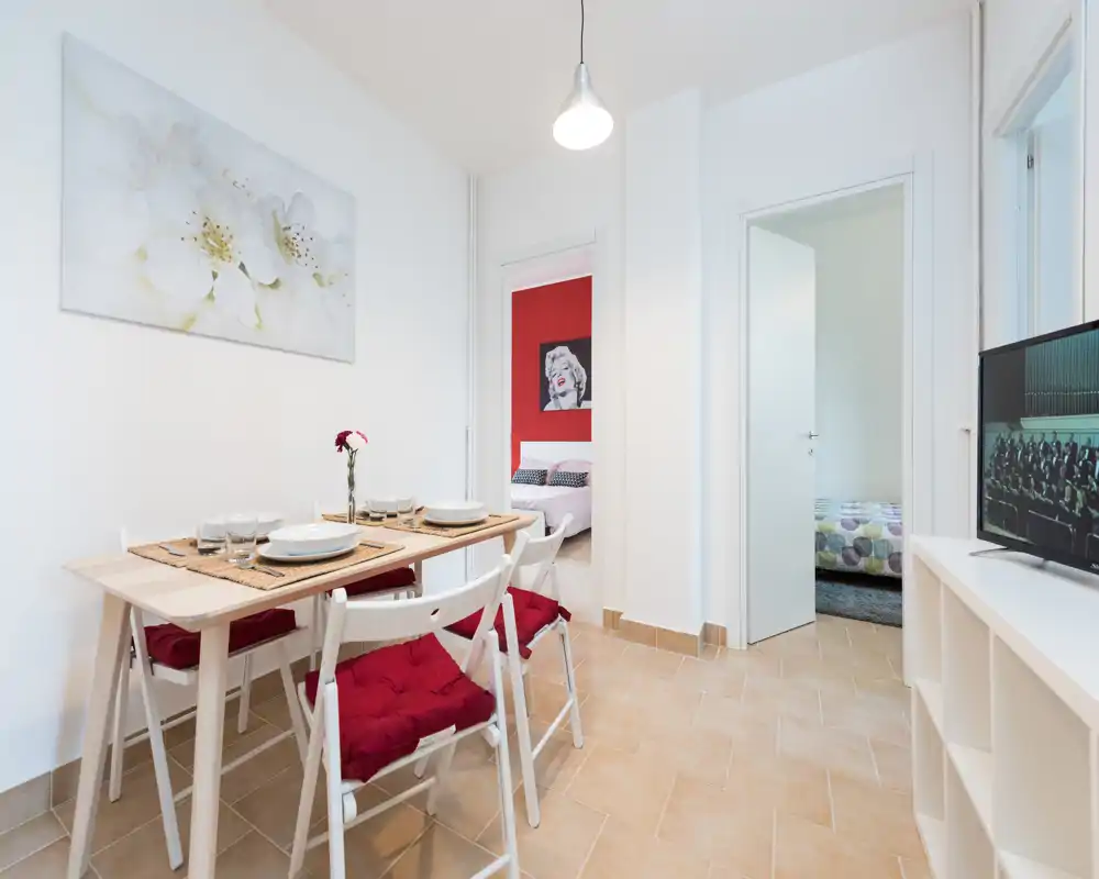 Bright, modern dining area with a set table for four, red seat cushions and minimalist decor; doorway views lead to bedrooms, creating a connected living layout.