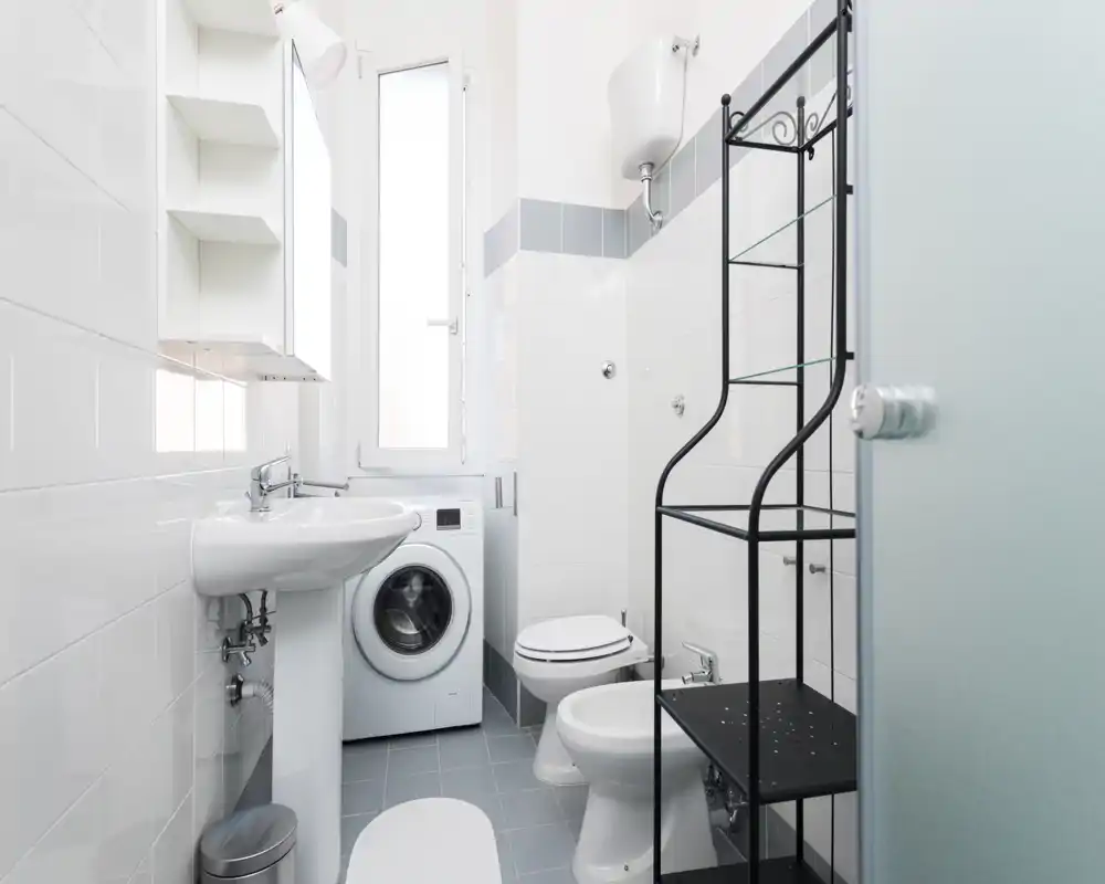 Clean, compact bathroom with white tiles, pedestal sink, toilet, bidet and a washing machine under a tall window; simple metal shelving provides storage.
