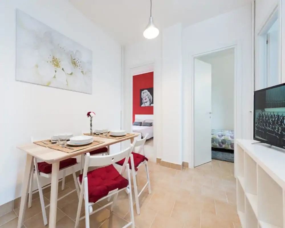 Bright, modern dining area with a wooden table set for four, red cushioned chairs and a floral centerpiece — ideal for casual meals in a clean, airy space.