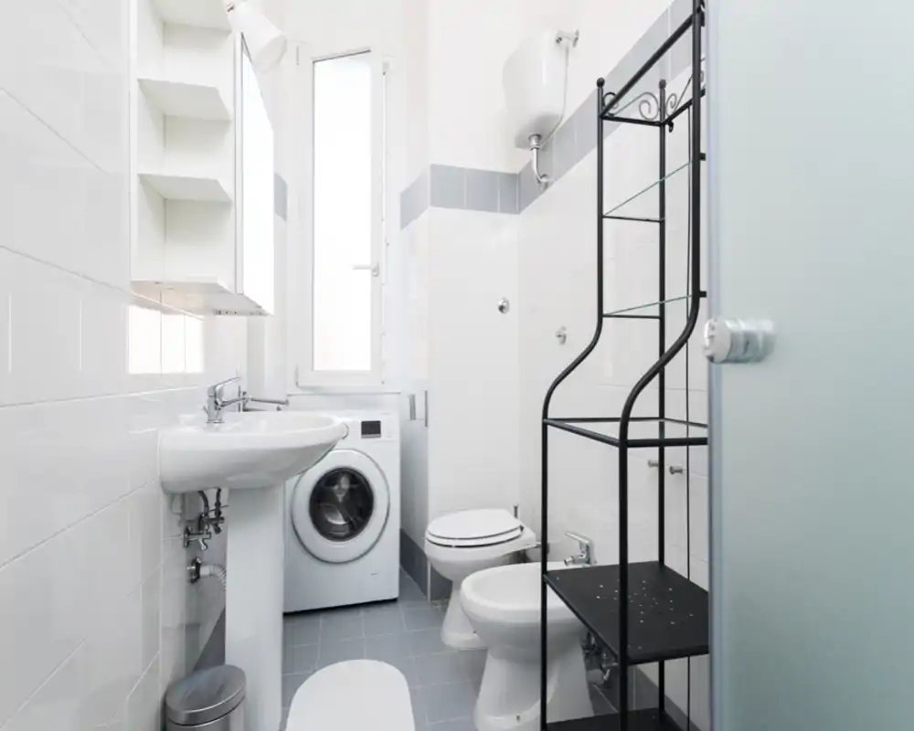 Bright, compact bathroom with white tiles, pedestal sink, toilet, bidet and a washing machine; neat shelving adds storage.