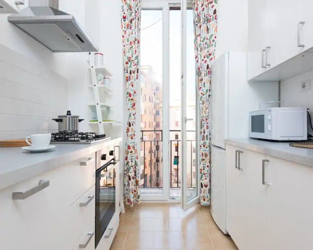 Bright, modern galley kitchen with white cabinets, built-in oven and gas hob, microwave and a tall window leading to a small balcony. Cheerful patterned curtains and tidy countertops create a fresh, welcoming space ideal for short-term rentals.