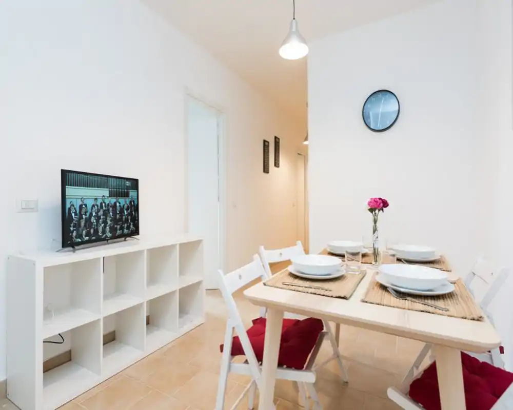 Bright, minimalist dining area with a wooden table set for four, white folding chairs with red cushions, and a small TV on a low shelving unit. The neutral walls and simple decor create a clean, inviting space ideal for small groups.