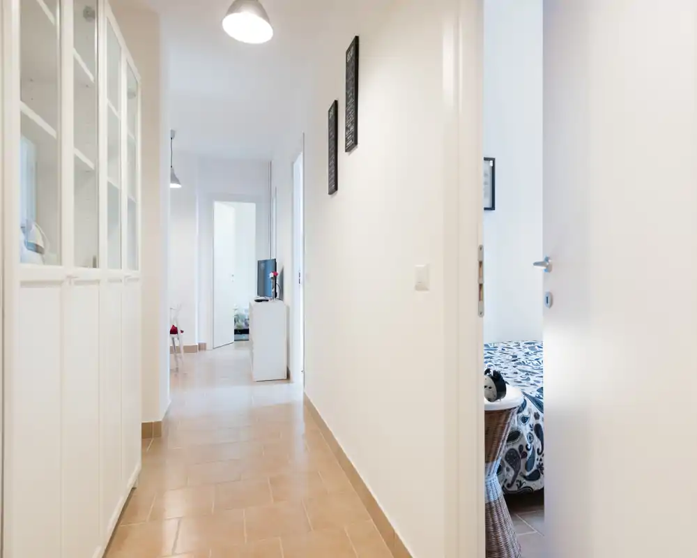 Bright, minimalist entrance corridor with clean white walls, tiled floor and wardrobe storage along the left side; doorway to a bedroom and further living area visible.