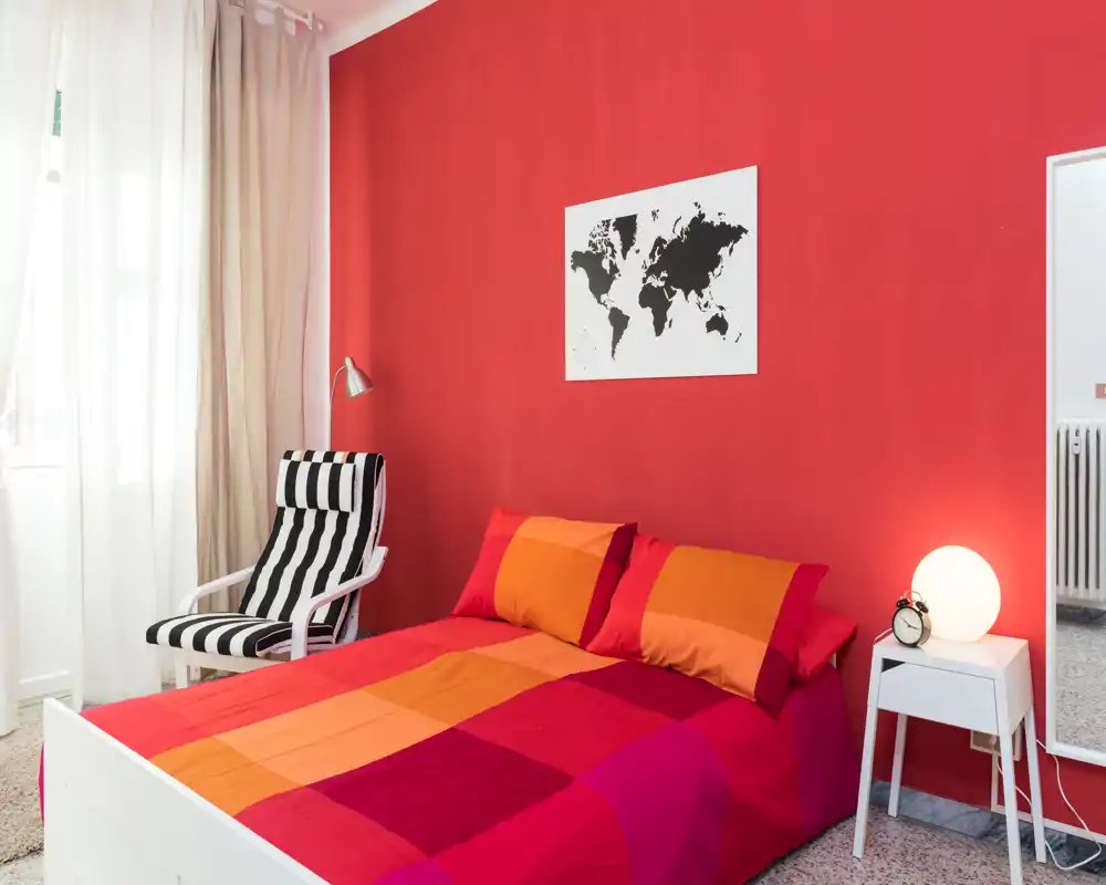 Camera da letto luminosa e accogliente con un letto matrimoniale colorato contro una parete rossa, comodino con lampada e una poltrona a righe vicino alla finestra.