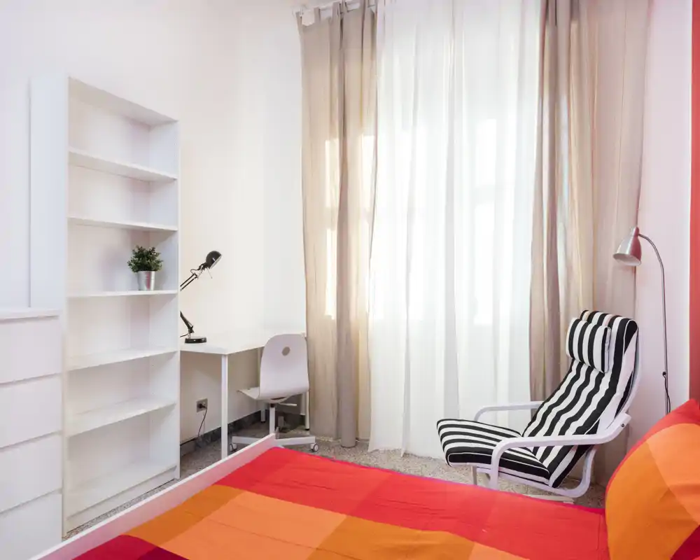 Camera da letto luminosa e moderna con copriletto colorato, poltrona a righe e una scrivania vicino alla finestra — perfetta per lavorare e rilassarsi.