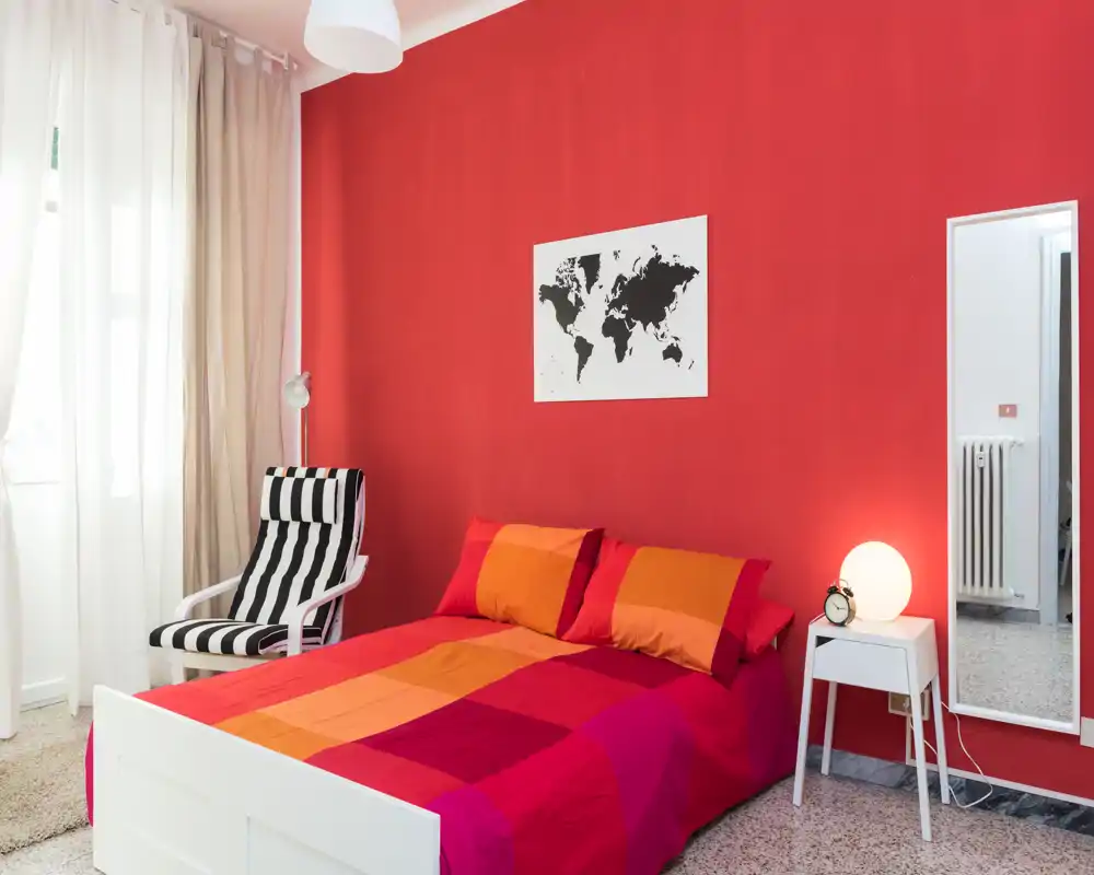 Camera da letto luminosa e colorata con letto matrimoniale con biancheria rossa-arancione, una poltrona a righe vicino alla finestra, comodino con lampada e specchio a figura intera — moderna e accogliente.