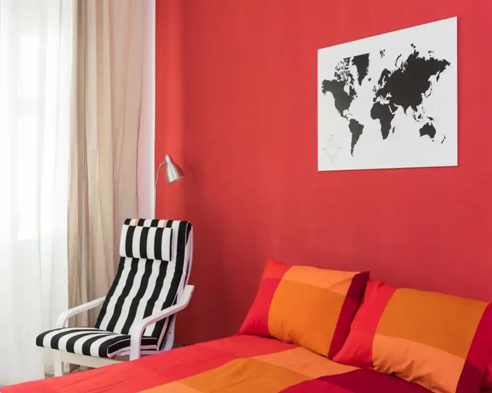 Camera da letto moderna e luminosa con parete rossa, poltrona a righe e copriletto arancione-rosso. La luce naturale attraverso le tende leggere crea un'atmosfera calda e accogliente, perfetta per annunci.