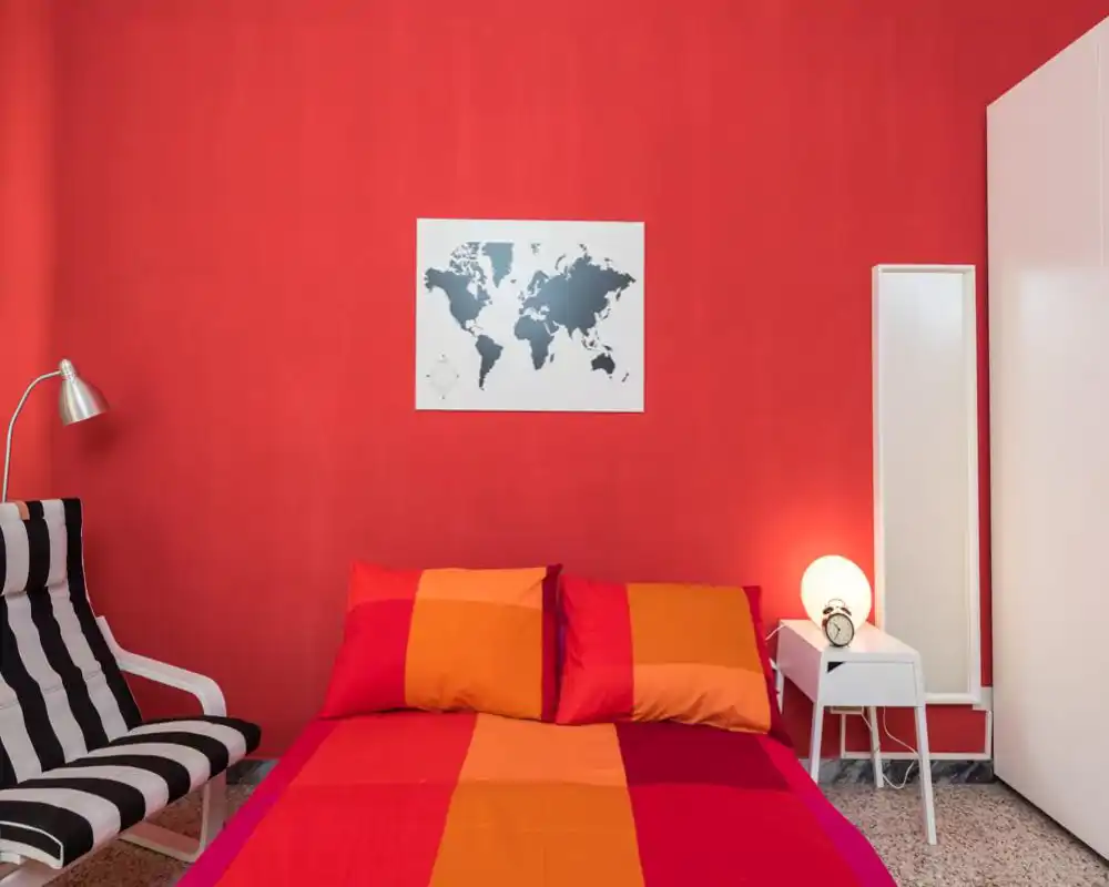 Camera da letto moderna e luminosa con un grande letto rosso-arancione contro una parete rossa, comodino con lampada e una poltrona a righe; colori vivaci e stile pulito rendono la stanza accogliente.