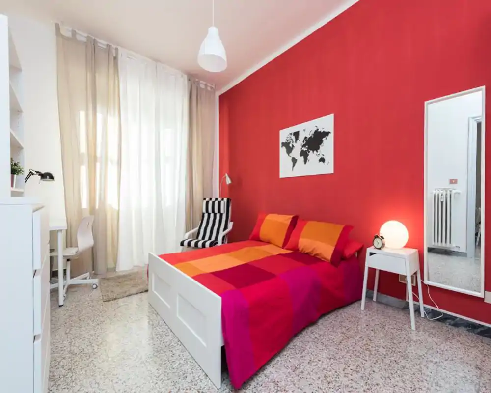 Camera da letto luminosa e moderna con struttura del letto bianca e biancheria colorata arancione-rossa su una parete accentuata di rosso vivo. Una poltrona a righe, comodino con lampada e specchio intero completano l'ambiente accogliente e di design.