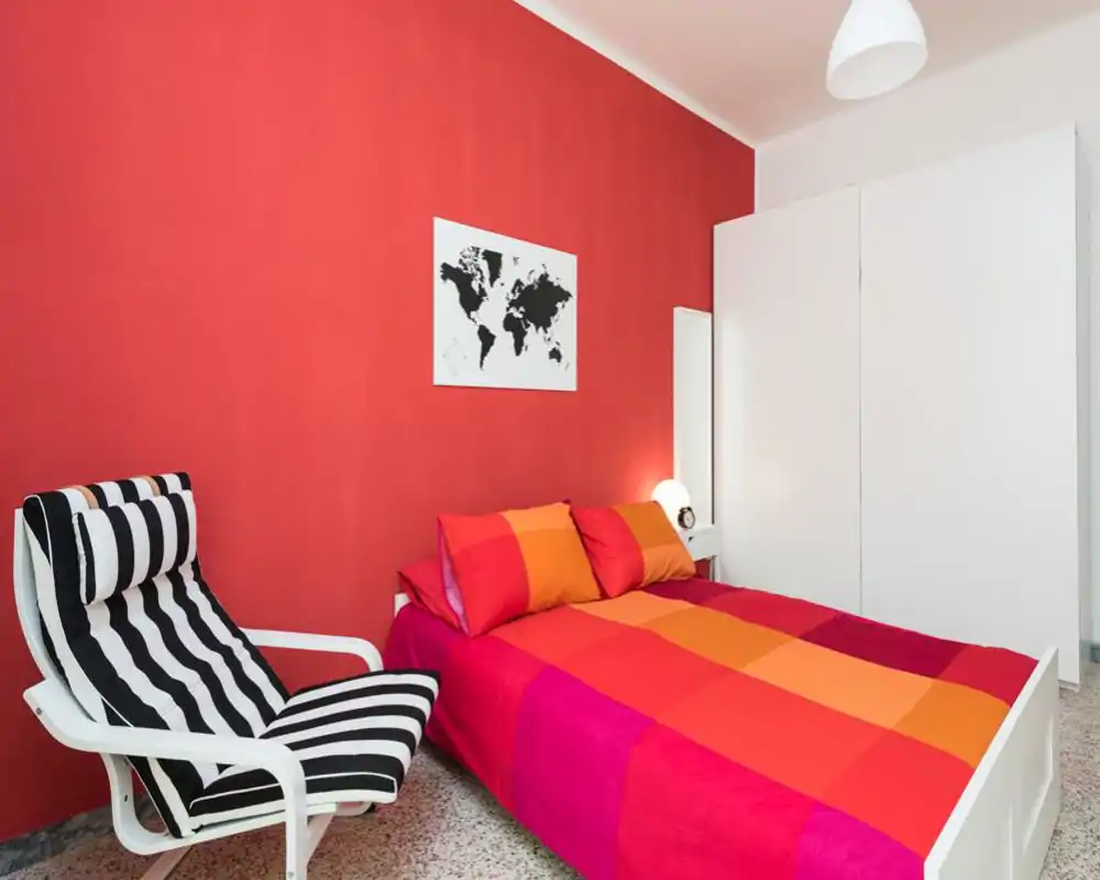 Camera da letto luminosa e vivace con letto matrimoniale con biancheria rossa e arancione, una poltrona a righe e un armadio bianco contro una parete d'accento rossa.