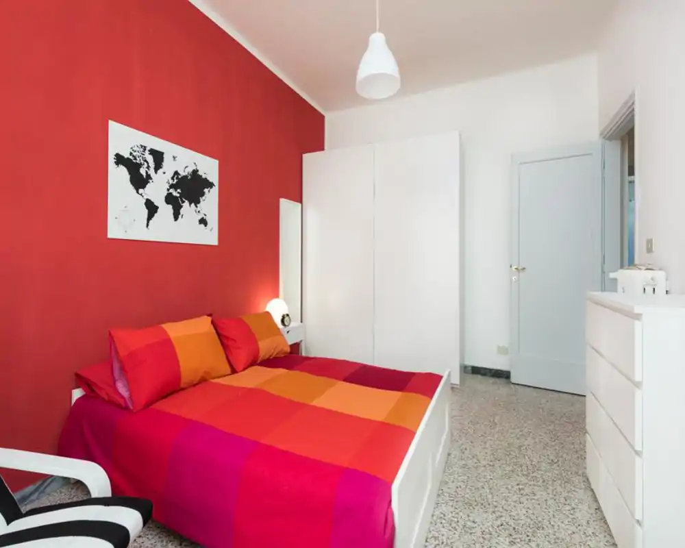 Camera da letto luminosa e contemporanea con letto matrimoniale colorato, armadio e comò bianchi. La parete rossa e la biancheria coordinata creano un ambiente moderno e accogliente, perfetto per annunci.