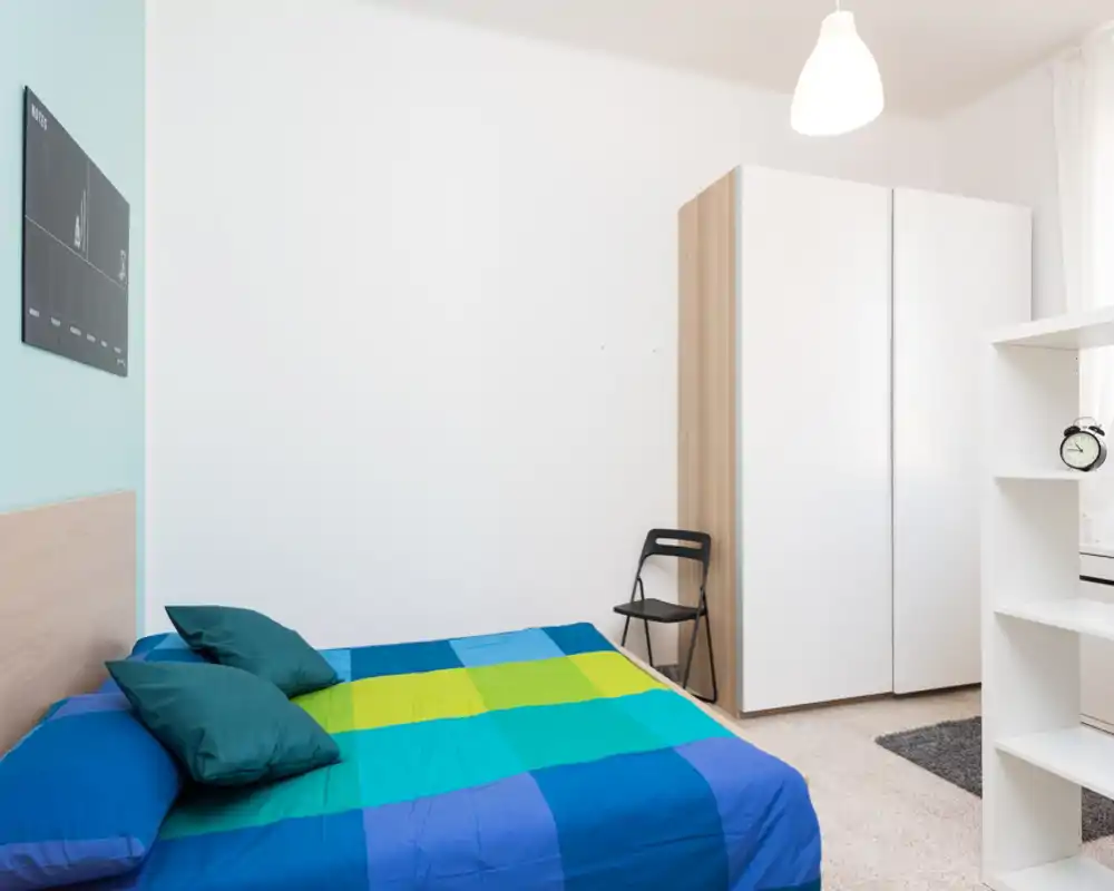 Bright, minimal bedroom with a double bed dressed in colorful blue-green bedding, a white wardrobe, a small chair and simple shelving — ideal for a modern rental listing.
