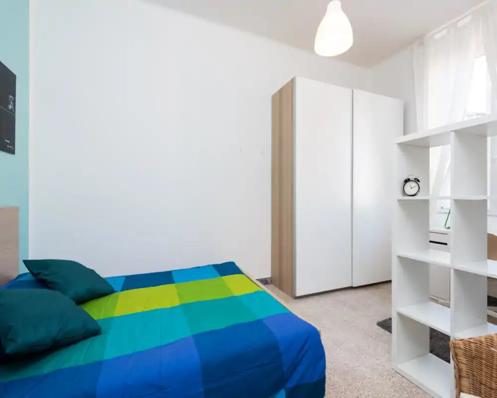 Bright, simply styled bedroom with a blue-and-green double bed, white wardrobe and shelving dividing the space. Natural light fills the room through a curtained window, creating a fresh, welcoming atmosphere ideal for rental photos.