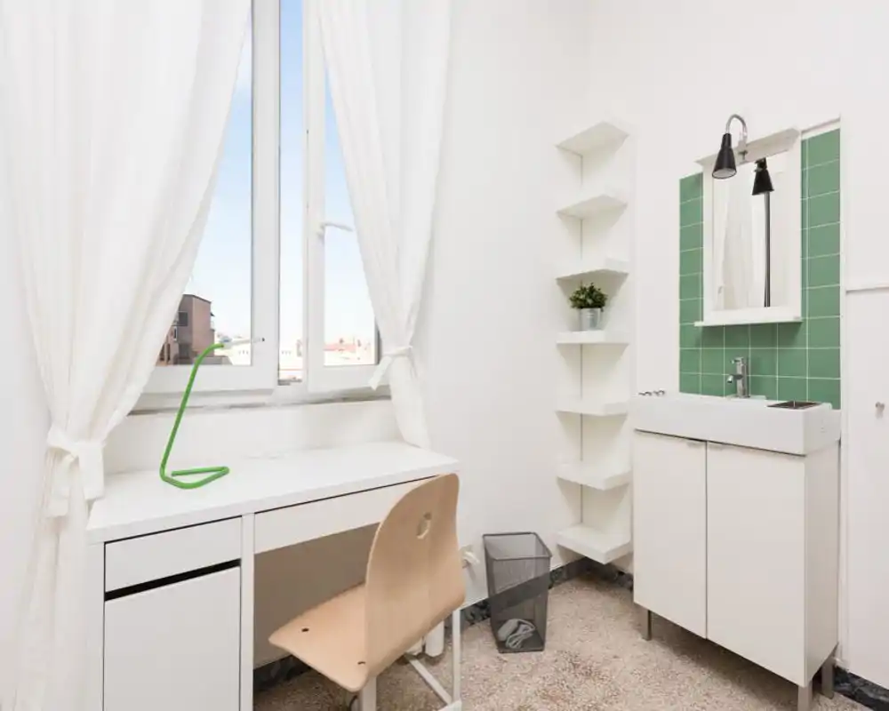 Bright, minimal room with a desk by the window, a small sink cabinet and open shelving — clean white decor and natural light make it feel fresh and functional.