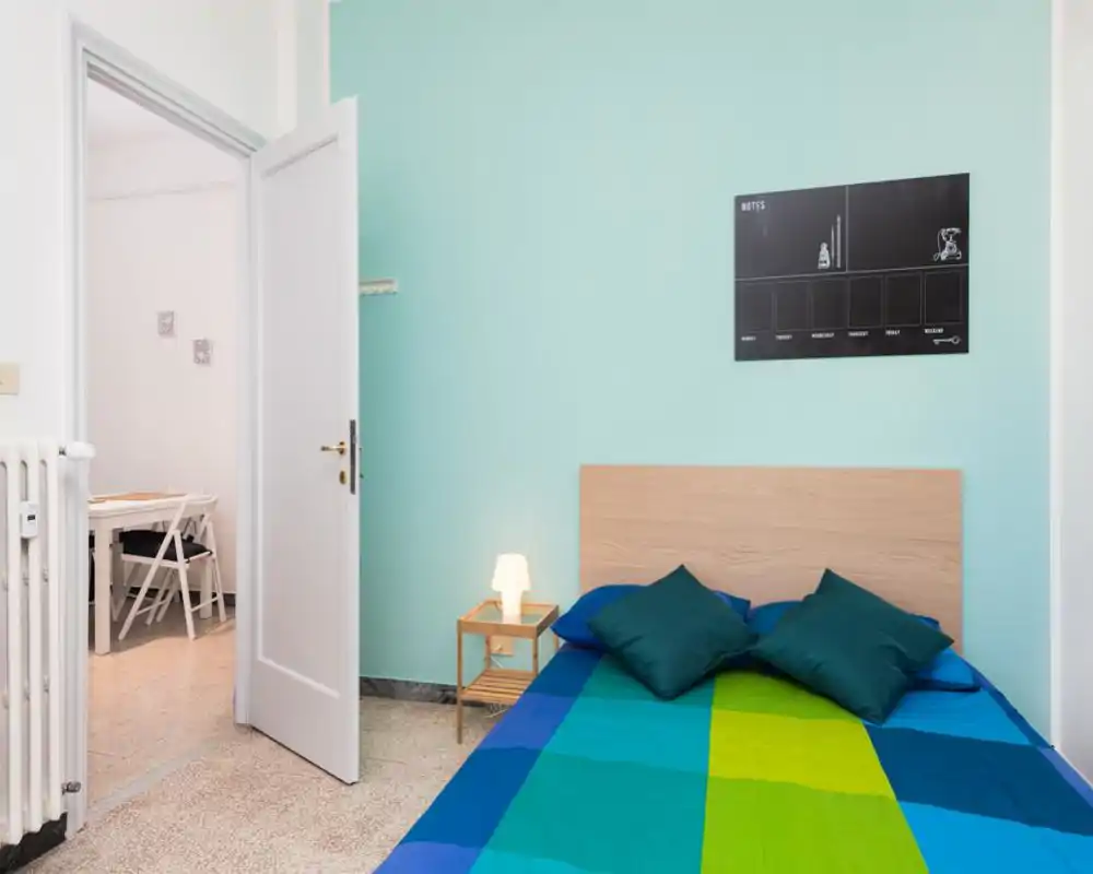 Bright, tidy bedroom with a double bed against a pale turquoise wall, wooden headboard, bedside table with lamp and decorative cushions; neat, modern styling ideal for rental photos.