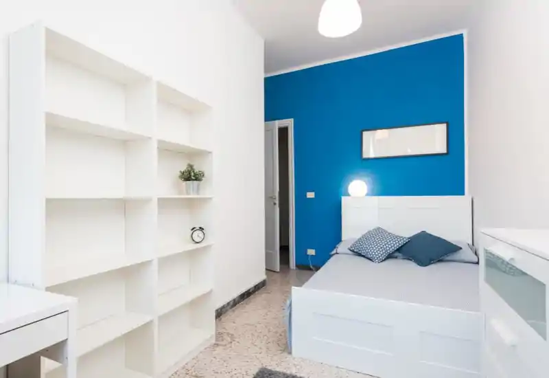 Camera da letto moderna e luminosa con letto bianco davanti a una parete blu acceso, cuscini decorativi e una lampada sul comodino. Scaffalatura minimal e comò completano lo spazio ordinato e accogliente.