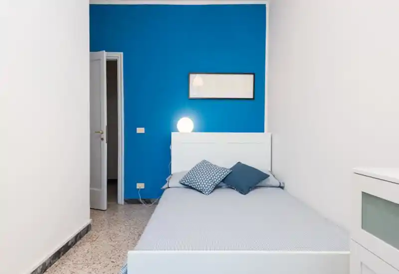 Camera da letto luminosa e ordinata con letto bianco su una parete blu acceso, cuscini decorativi e una lampada da comodino che creano un’atmosfera accogliente.