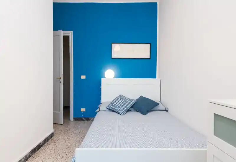 Camera da letto luminosa e moderna con un letto in legno bianco contro una parete blu, cuscini decorativi e una lampada da comodino che creano un aspetto pulito e confortevole.