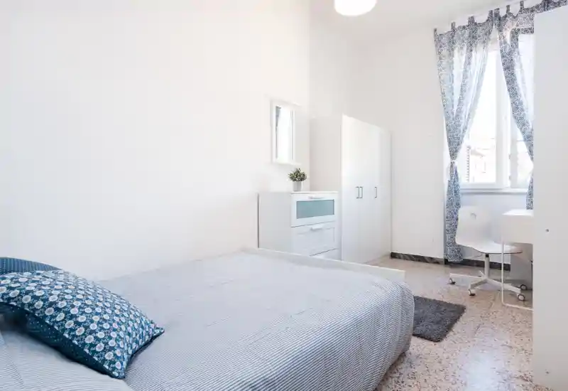 Camera da letto luminosa e ariosa con letto matrimoniale, cuscini coordinati e tende con motivo; armadio, comò e scrivania vicino alla finestra per un soggiorno confortevole.