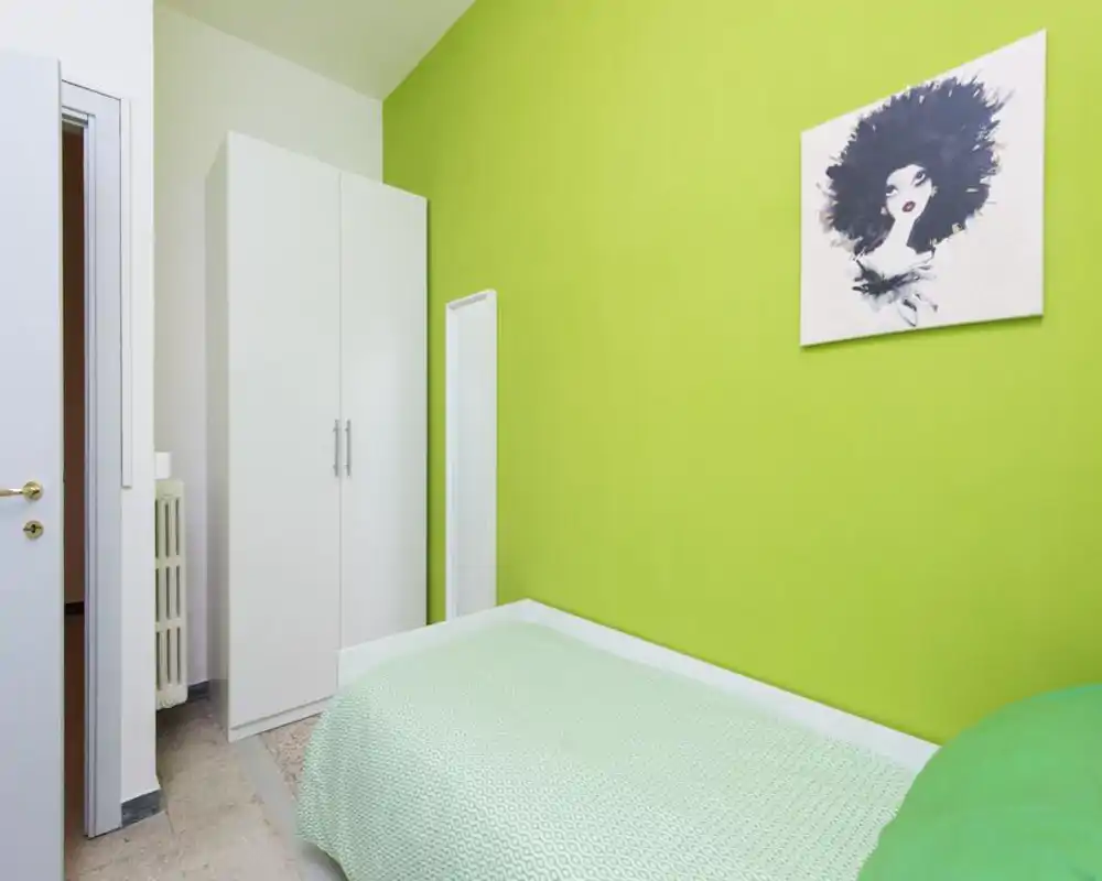 Habitación pequeña y ordenada con cama individual junto a una pared verde brillante, armario blanco y un espejo vertical; luz limpia y decoración minimalista.