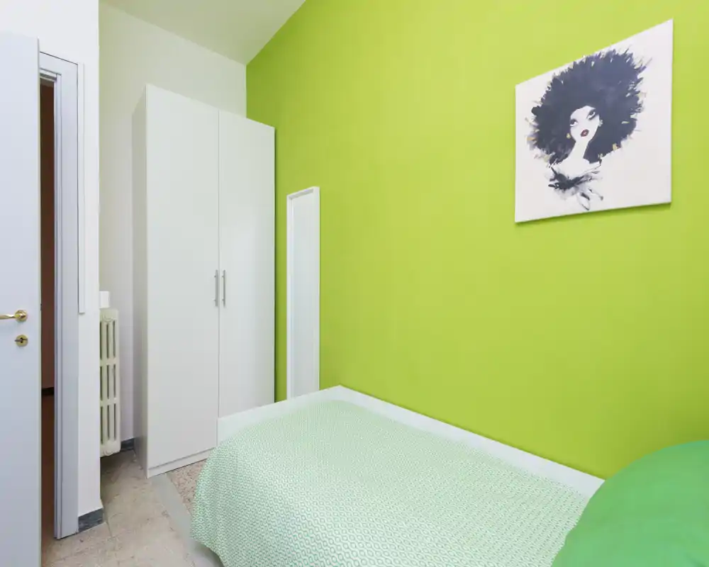 Dormitorio pequeño y luminoso con cama individual junto a una pared verde lima, armario blanco y cuadro decorativo. Estilo limpio y moderno con buena luz, ideal para estancias cortas y acogedoras.