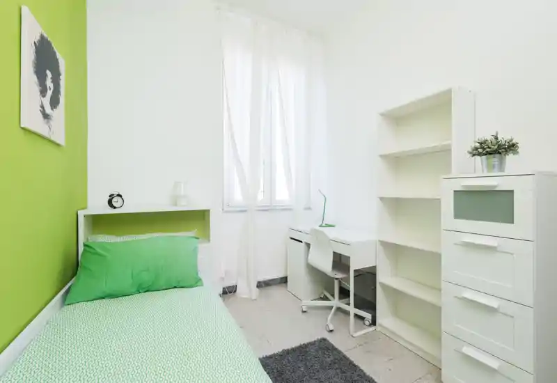 Camera singola compatta e luminosa con letto in verde, scrivania bianca e scaffalatura — ideale per uno studente o soggiorni brevi. La stanza è ordinata, ben illuminata dalla finestra centrale e arredata con tessili coordinati.
