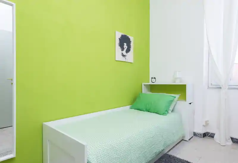 Dormitorio individual compacto con cama de estructura blanca, pared verde y ropa de cama a juego; luz natural intensa por la ventana y mesilla minimalista.