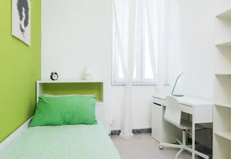 Camera da letto compatta e luminosa con letto singolo con biancheria verde, mensola con lampada e sveglia, e scrivania bianca vicino alla finestra — pulita e ben illuminata.