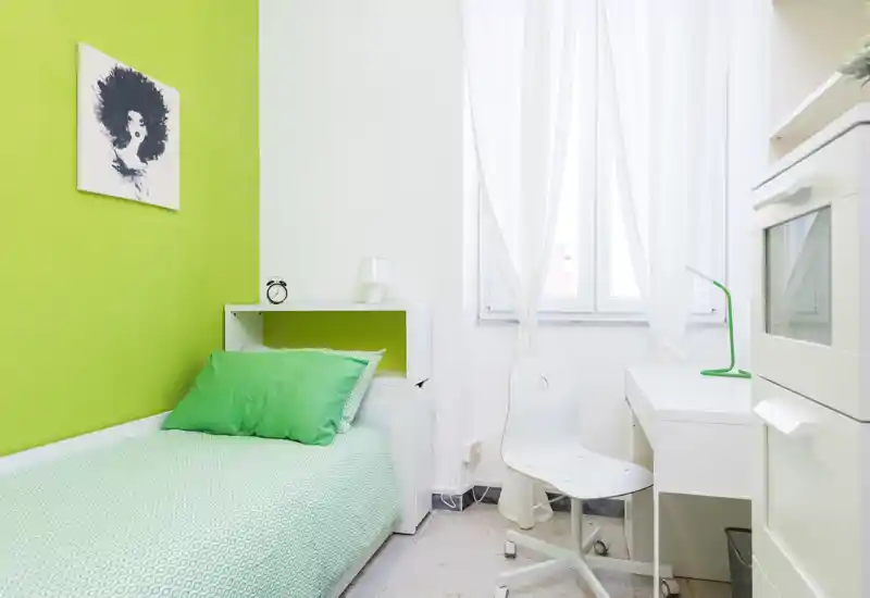 Dormitorio individual luminoso con pared verde, cama bien hecha y un escritorio blanco junto a la ventana — ideal para estudiante o una sola persona.