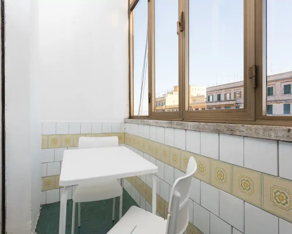 Angolo pranzo piccolo con tavolo e sedie bianche accanto a una grande finestra con vista sulla città; piastrelle pulite e luce naturale creano un'area pranzo semplice e funzionale.