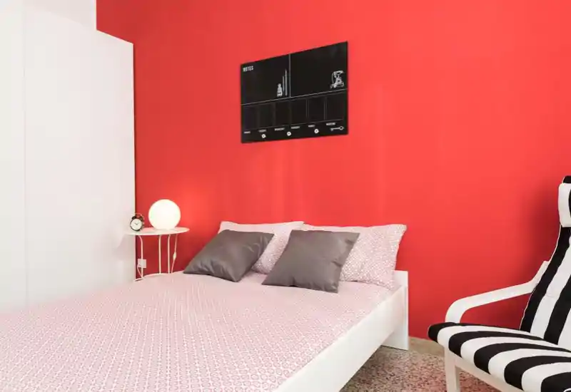 Camera da letto moderna e luminosa con letto a piattaforma bianco contro una parete rossa, biancheria rosa a motivo, due cuscini grigi e un comodino con lampada sferica.